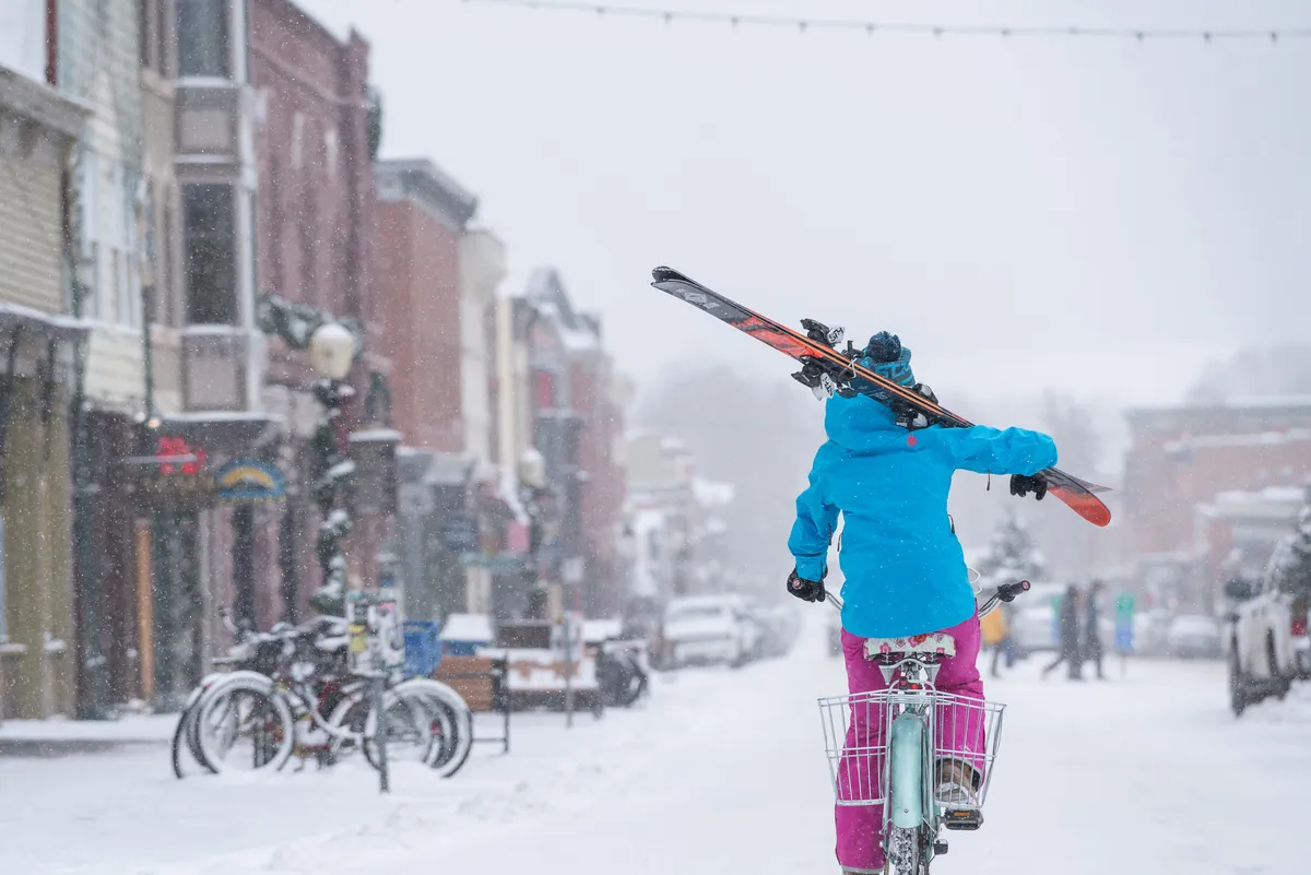 An active street scene with a person skiing with skis attached to a bicycle on a snow-covered road, surrounded by historic buildings and bicycles. The image conveys a sense of fun, adventure, and the unique character of the local area.