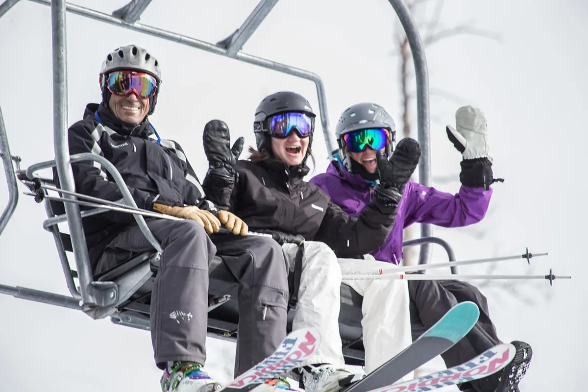Three skiers ride a chairlift over a snow-blanketed landscape, clad in colorful ski gear, with frosted trees and bright sky setting the winter adventure mood. The image evokes the thrill and camaraderie of hitting the slopes, with Telluride's alpine environment on full display.