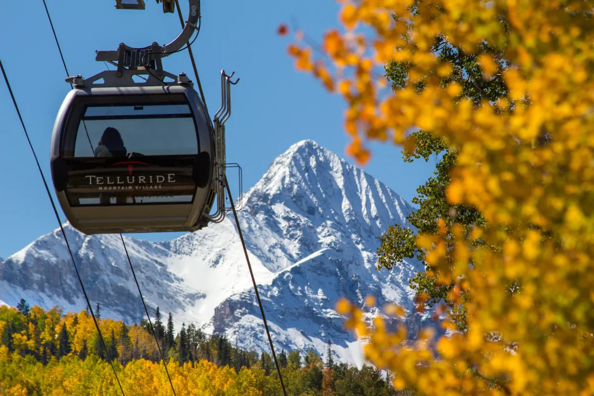 This image highlights a gondola ascending towards a snow-capped mountain, set against vibrant autumn foliage. The scene captures the out-of-door, adventurous atmosphere of Telluride, with the gondola hinting at easy access to both natural wonders and local amenities.