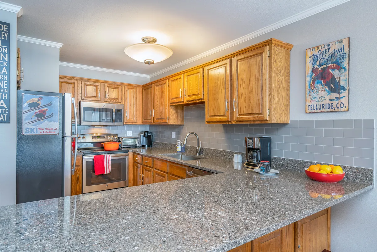 A spacious kitchen with a granite countertop, wooden cabinets, modern appliances, and a bowl of fruit on display. The kitchen is clearly designed for group meal prep and entertaining, inviting guests to gather around while food is being prepared.
