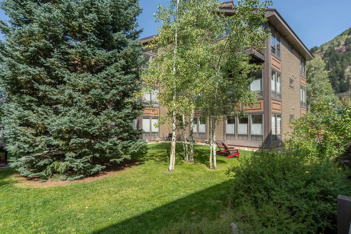 The lawn outside Boomerang Lodge features tall evergreen and aspen trees, red chairs for lounging, and a peaceful building facade. The space is inviting for post-adventure relaxation or casual gatherings, with hints of both summer and winter charm.