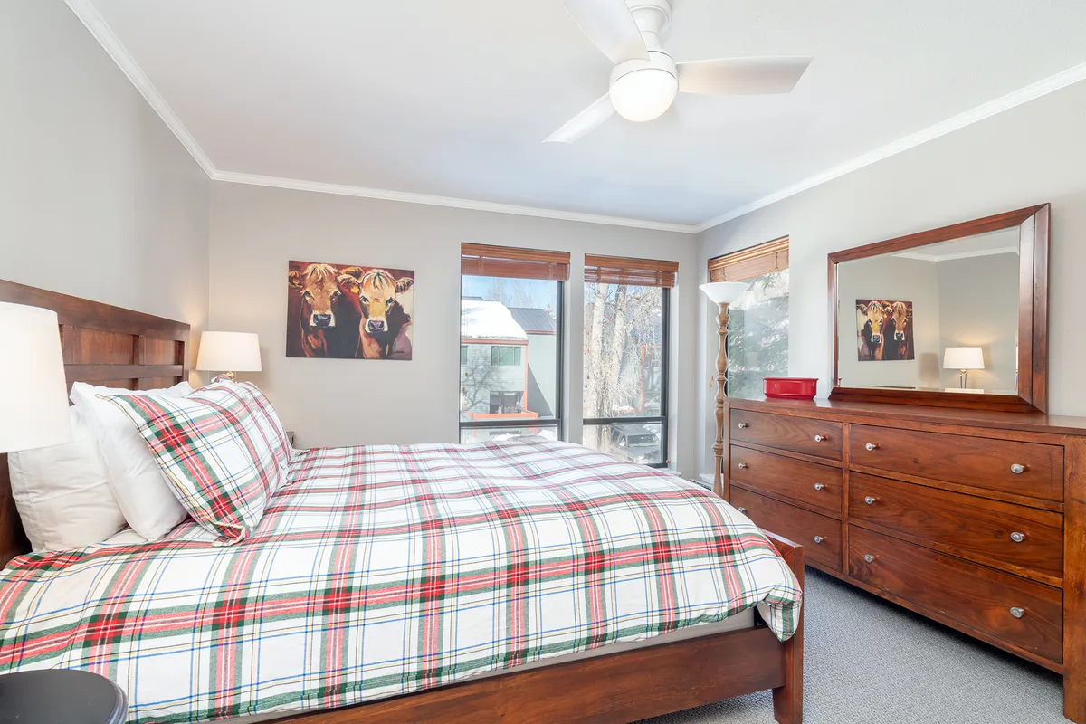 A sunlit bedroom featuring a plush queen bed dressed in plaid bedding, a wooden dresser, a large window offering ample natural light, and a playful cow artwork above the headboard. The space feels warm, comfortable, and perfect for relaxation after a day outdoors.