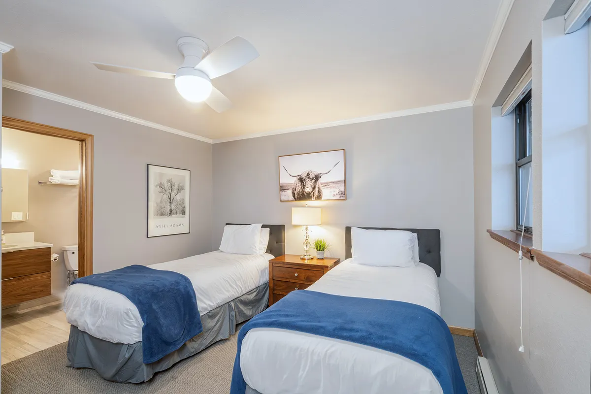 A cozy bedroom with two twin beds separated by a nightstand with a lamp. Framed cow artwork adds a whimsical touch, and the room is softly lit, creating a peaceful environment suited for both kids and adults. The flexible arrangement reflects the twin/king conversion option.