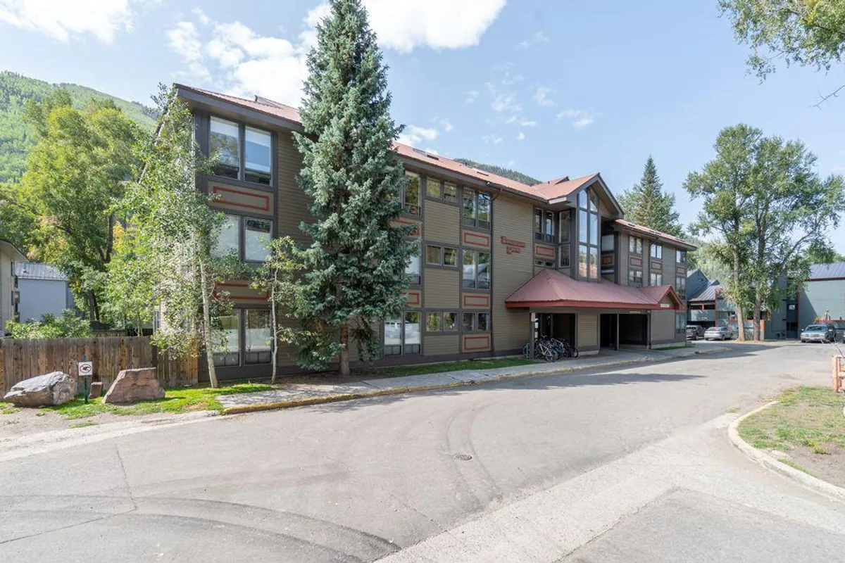A clear view of the multi-story building with green trees and a visible parking area in the foreground. Bicycles near the entrance hint at active, community-focused living. The parking lot is easily identifiable, suggesting convenience and security for residents.