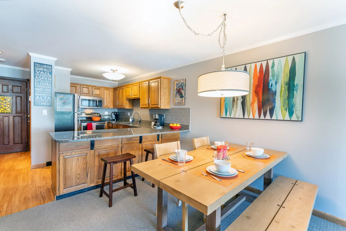 A warmly lit dining table is beautifully set, positioned near a kitchen countertop and framed by colorful wall art and wooden cabinetry. The space is alive with the spirit of shared meals, celebrations, and casual gatherings, inviting everyone to come together for food and conversation.