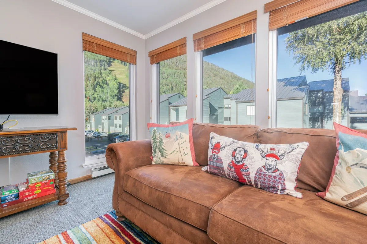 A comfortable brown couch adorned with colorful throw pillows sits beside large windows that bring in natural light and showcase scenic views. The flat screen TV is visible, ideal for movie nights or game sessions. The layout encourages relaxation and togetherness, perfectly suited for cozy evenings with family or friends.