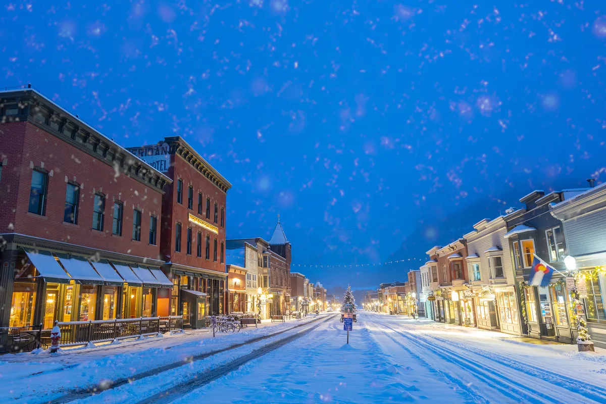 A charming town street lined with historic buildings, blanketed in snow and adorned with festive string lights and a decorated Christmas tree. The scene radiates warmth, community, and a welcoming atmosphere—ideal for exploring, shopping, or dining as a family.