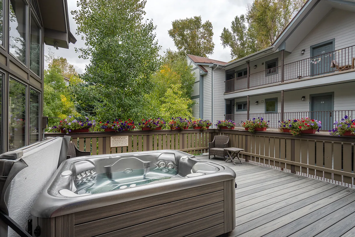 A serene wooden deck houses a bubbling hot tub surrounded by flower boxes, set against a backdrop of natural beauty. The inviting setup is designed for relaxation and socializing in the fresh mountain air, emphasizing the resort-like amenities available to residents.