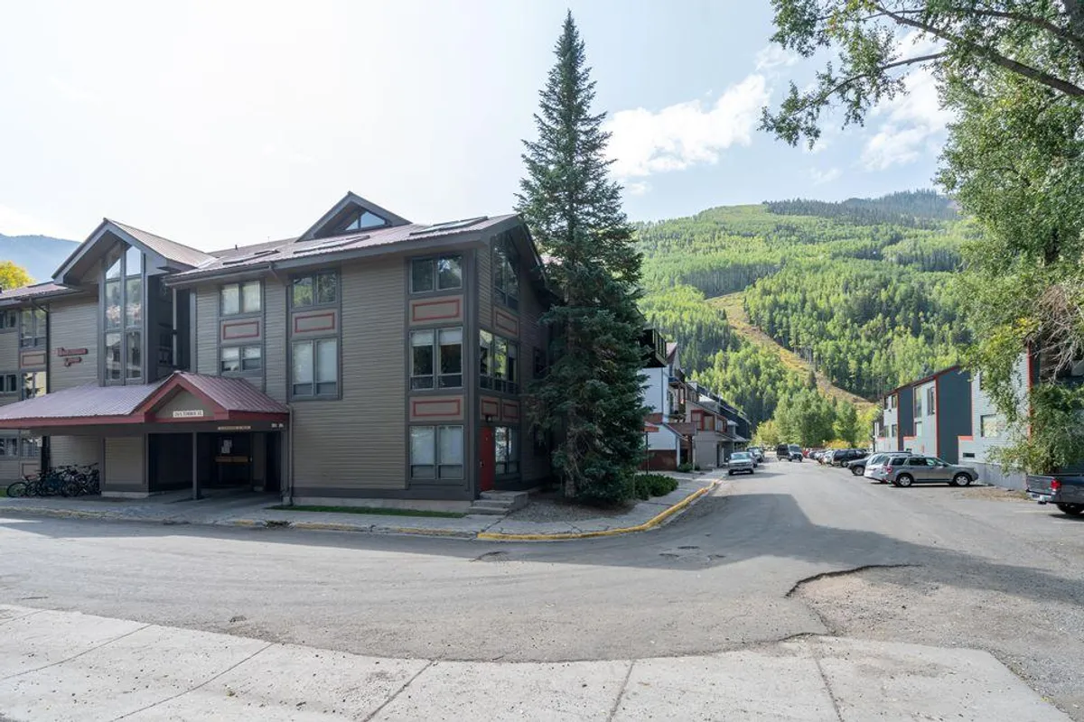 The building exterior framed by lush trees and a striking mountain backdrop, highlighting the property’s setting within Telluride’s scenic landscape. Sidewalks and open spaces suggest outdoor walkability and proximity to natural beauty.