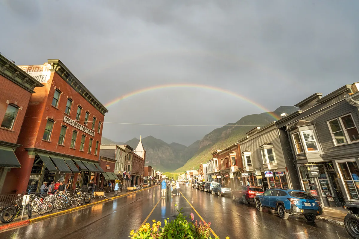 A vibrant street view featuring historic storefronts, a beautiful rainbow arching overhead, and wet pavement reflecting the town's colorful charm. Mountains rise in the background, reinforcing Telluride’s picturesque setting and walkable, community-oriented vibe.