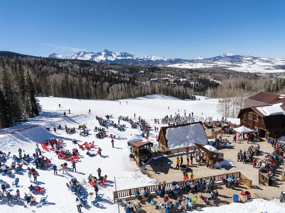 A lively exterior scene showing a crowd gathered at the base of snow-covered slopes, set against a beautiful mountain backdrop and rustic wooden buildings. People are seen relaxing, socializing, and enjoying the outdoors together.