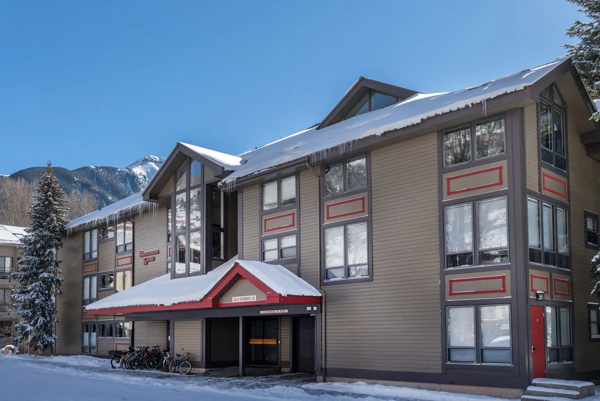 This exterior image features Boomerang Lodge with snow-covered roofs, large windows, and red architectural accents. Bicycles are parked outside, suggesting year-round access and an active, social atmosphere. The lodge is nestled amidst scenic mountain views, creating an inviting base for group gatherings.
