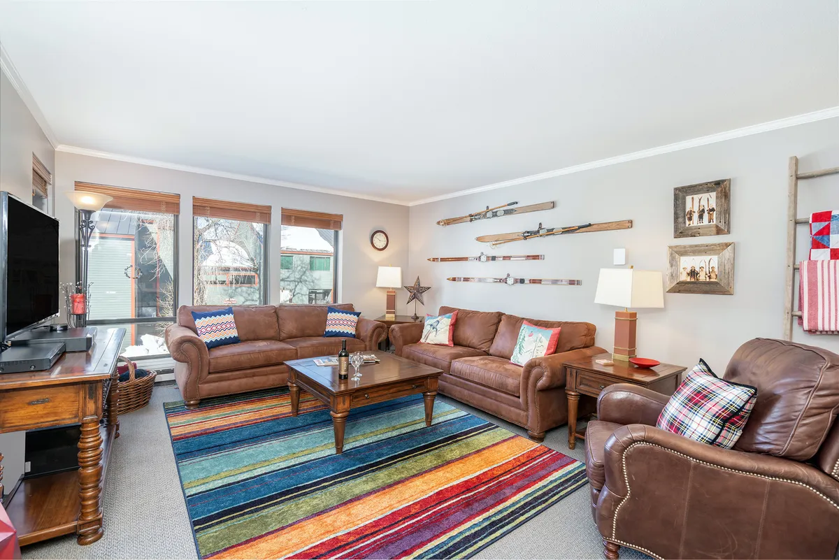 The image showcases a warm and inviting living area featuring plush leather sofas arranged around a vibrant, colorful rug and a stylish coffee table. Decorative wall elements add character, while the space is bathed in soft, natural light. This open layout is perfect for relaxing, socializing, or enjoying a cozy movie night—ideal for families or groups after a day of Telluride adventure.